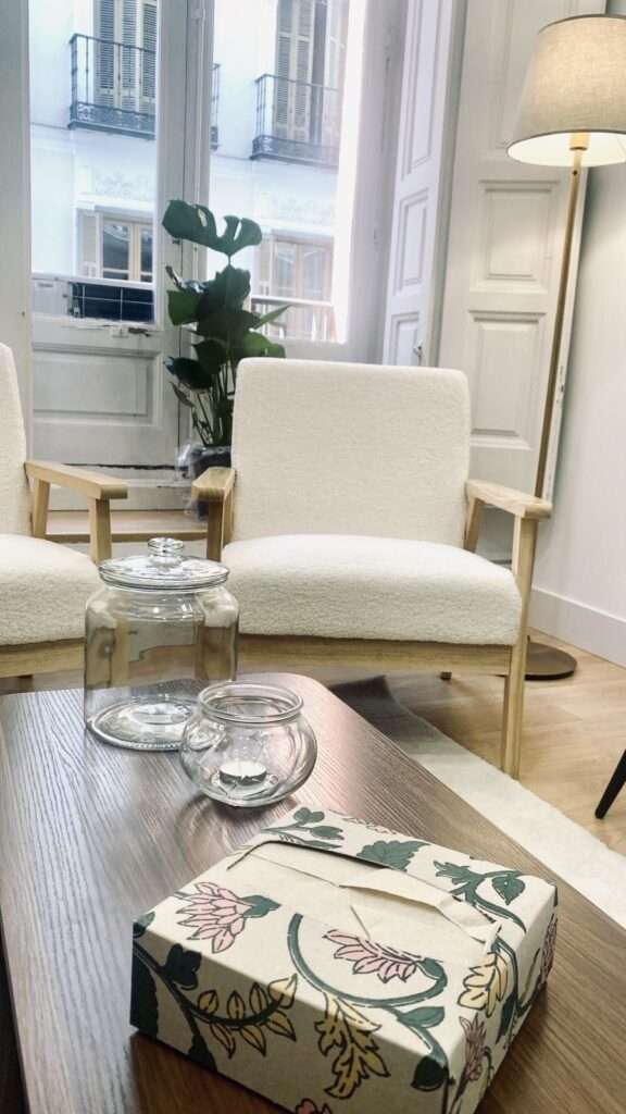 mesa con pañuelos, una vela y un jarron; de fondo se ven dos butacas y una ventana